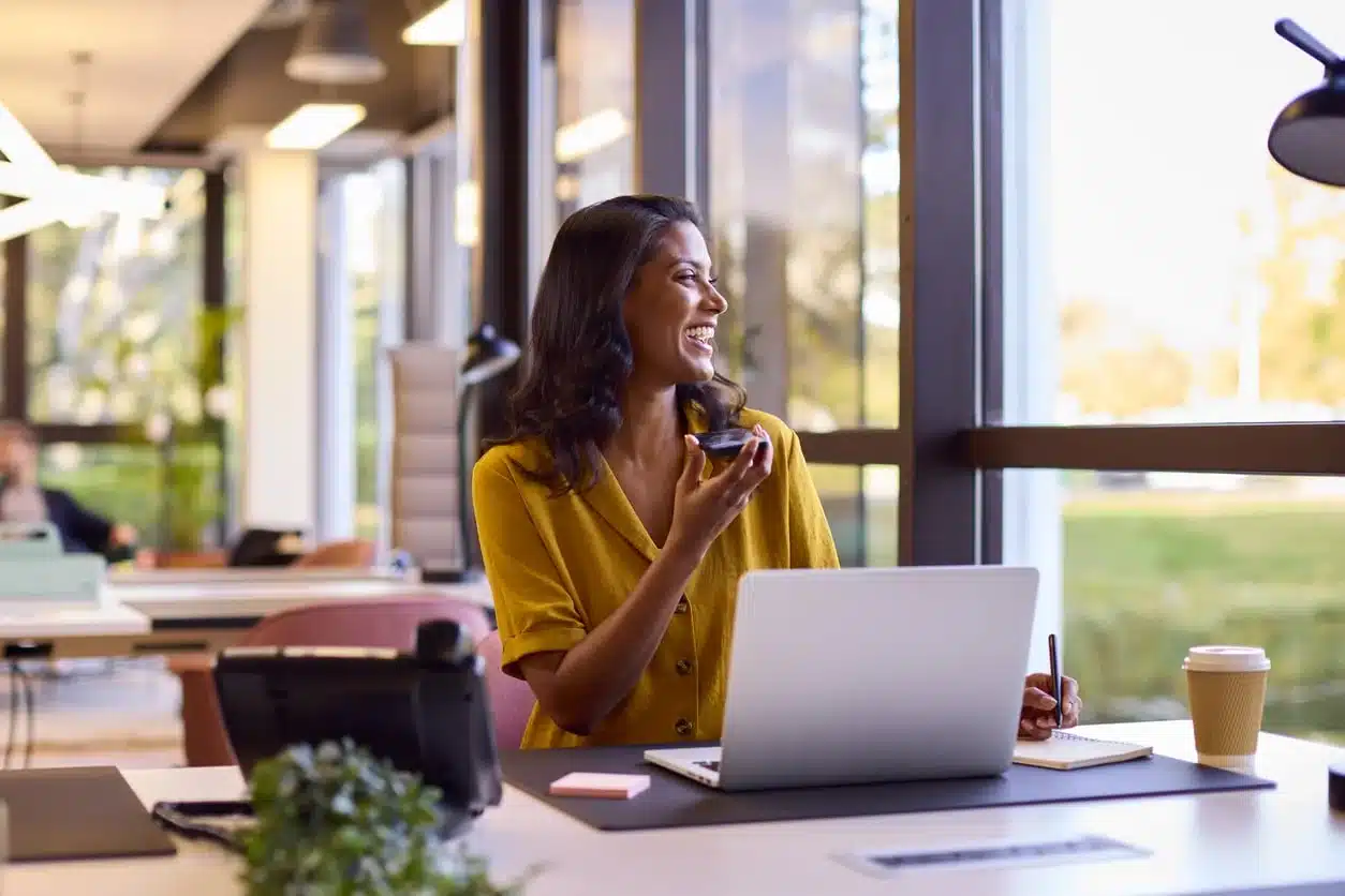 Woman in a yellow blouse smiling on a phone call at a modern coworking space with a laptop and notebook, working abroad while claiming the Foreign Earned Income Exclusion.