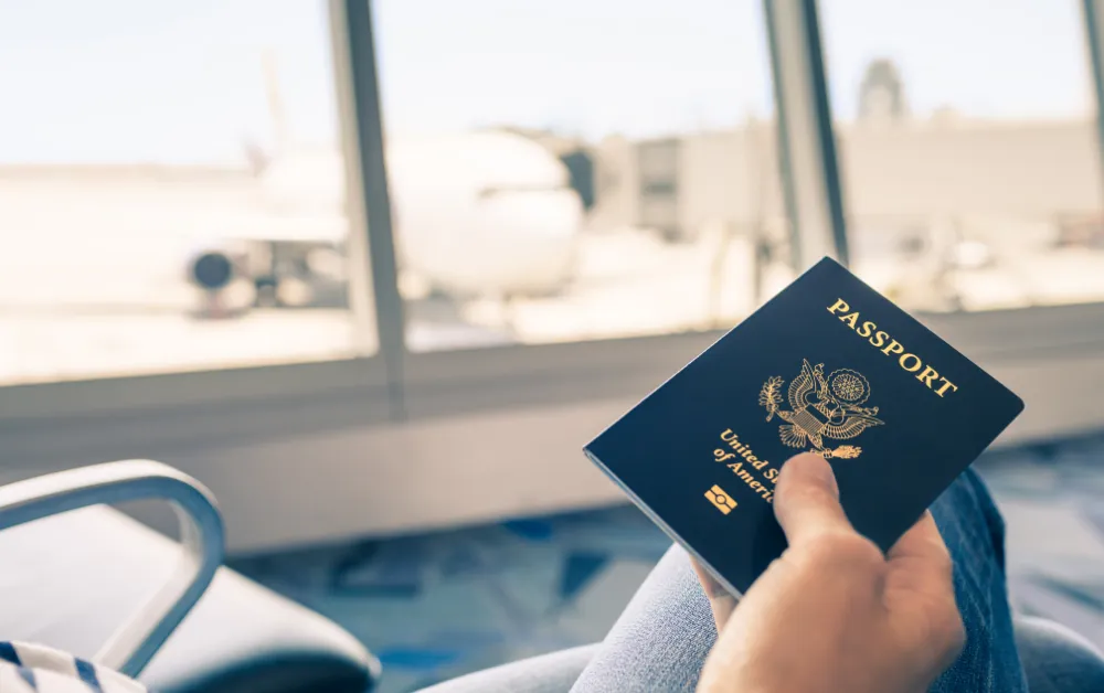 Hand holding a U.S. passport at an airport gate with a plane visible through the window, preparing to leave the country.